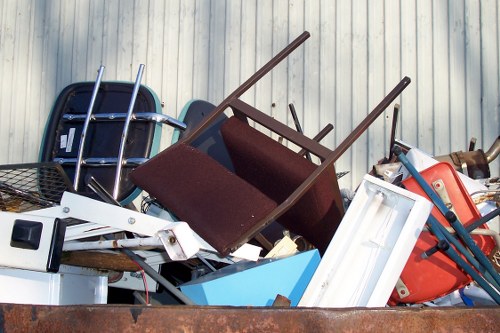 A large pile of assorted rubbish and discarded items stacked outdoors on a concrete surface. The collection includes various furniture pieces such as a brown fabric-upholstered armchair with wooden legs, a white plastic frame possibly from a small appliance or window, and a blue plastic bin. There are also metal objects, including a blue-handled wheelbarrow or cart, and other miscellaneous debris. The background features a vertically ribbed, light-colored metal fence, indicating an industrial or commercial premises near Earls Court, London. The scene is illuminated by natural daylight, highlighting the textures and colors of the different materials. This image visually represents waste awaiting collection or removal by a professional rubbish removal service like Commercial Waste Earls Court, operating within the local area, highlighting the typical types of refuse managed in urban environments within postcode regions such as SW5 or SW10.