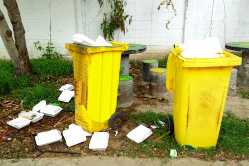 The image depicts two large, yellow plastic wheelie bins used for rubbish disposal, positioned outdoors on a grassy and paved area. The bins are situated in front of a white painted brick wall, with a tree trunk visible on the left side of the image and some green foliage or climbing plants hanging over the wall. One bin is slightly to the left and the other to the right, both with open lids revealing some white waste inside. Several white foam food containers and a few small pieces of trash are scattered on the ground around the bins, indicating improper disposal outside the bins. The environment appears to be part of a residential or small commercial area, potentially in a town setting like Earls Court. The overall scene highlights the need for rubbish collection services, such as those provided by Commercial Waste Earls Court, and demonstrates typical waste management issues related to littering and bin usage in this context.