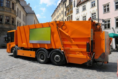 A large, orange commercial waste collection vehicle parked on a cobbled street within a town environment, with multi-story Victorian and Georgian-style buildings in the background. The truck features a rectangular, enclosed rear compartment designed for rubbish collection, with visible hydraulic mechanisms and safety barriers. Its vibrant orange colour contrasts with the light grey cobblestone surface and the surrounding architecture. The vehicle's side panel includes a reflective green and silver safety strip, and the rear section has a sealed, sloped lid and load area, indicating it is ready for waste collection or disposal. The area appears clean and well-maintained, typical of municipal waste management operations, with no visible debris on the street. The scene is set during daylight, with natural light illuminating the details of the waste collection lorry and the surrounding buildings, subtly reflecting the typical rubbish removal service in Earls Court, London, with a domain relevance to commercial waste management in central London.