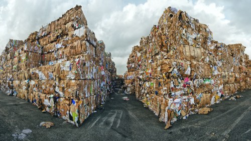 A large outdoor waste storage area featuring two extensive stacks of compressed cardboard and paper waste bales, arranged in a rectangular formation with a narrow pathway between them. The bales are tightly packed and stacked high, displaying a rough, textured surface with visible layers of corrugated cardboard, flattened boxes, and printed paper, showing various shades of brown, beige, and white. The environment appears to be an industrial or commercial site with a paved surface underneath and a cloudy sky overhead. The setting suggests a site suitable for rubbish removal services, such as those offered by Commercial Waste Earls Court, serving local businesses in the UK postal code area near Earl's Court, London.