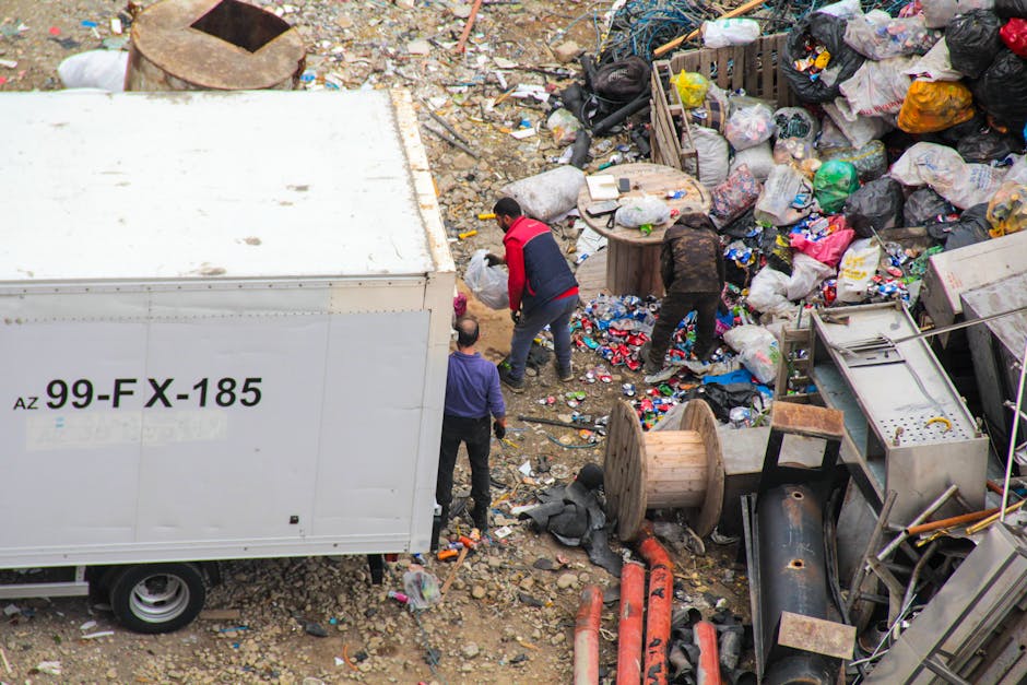 A large outdoor waste collection point with multiple bins and overflowing rubbish bags positioned on a paved area beside a parked silver car. The bins include a grey mixed waste container with a blue 