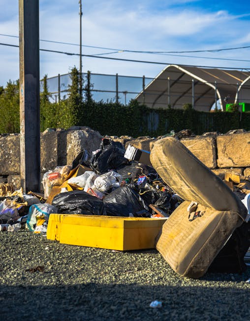 A large pile of mixed waste is situated on an asphalt surface in an outdoor area, with predominantly black plastic rubbish bags, some white and clear bags, and loose debris, including paper and small 